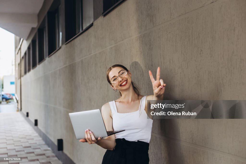 Young woman showing v-sign and holding laptop.