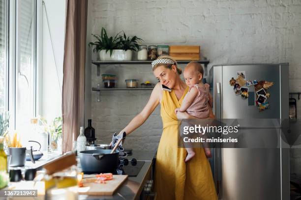 beautiful woman holding a baby while talking on the phone and cooking at home - busy kitchen stock pictures, royalty-free photos & images