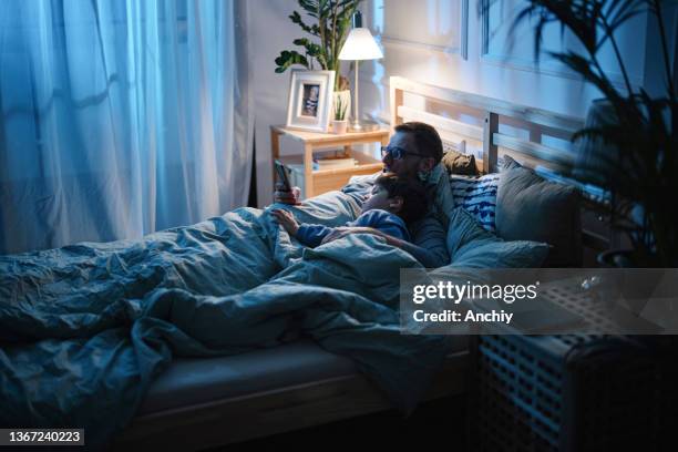 adorable little boy and his father using a smartphone together at night in bed - manly room stock pictures, royalty-free photos & images