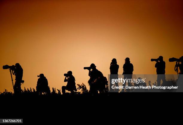 Photographed in silhouette, back lit, sunset light, amateur photographers use their cameras, telephoto lenses, and monopods while photographing the...