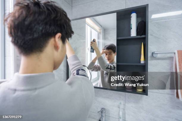 a young male looks in the mirror and sprays hairspray on his hair - man combing hair stock pictures, royalty-free photos & images