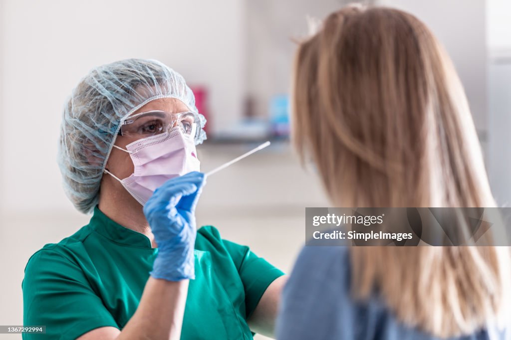 Well-protected medical staff performs swab test for Covid-19 to a young blonde female patient.