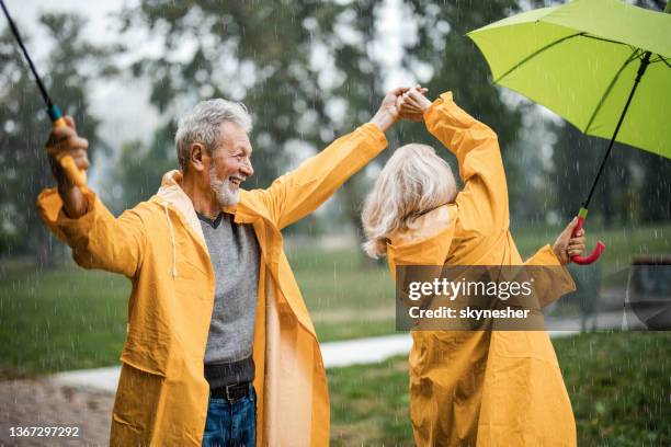 carefree senior couple in raincoats dancing on a rainy day. - yellow umbrella stock pictures, royalty-free photos & images