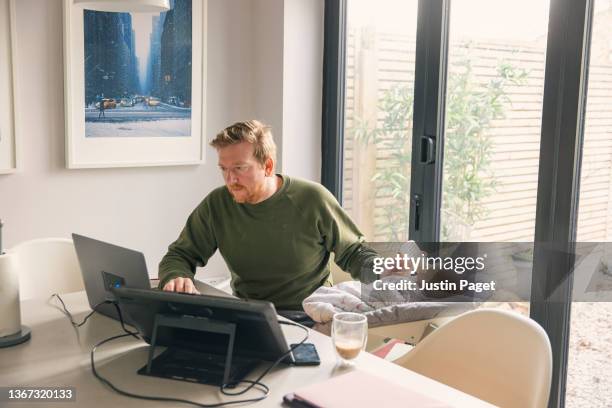 man working from home dining table whilst feeding his baby daughter - multi tasking foto e immagini stock
