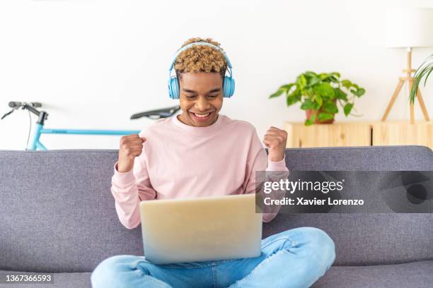 excited teenage boy celebrating good news while using a laptop computer sitting on a sofa at home. - excitement laptop stock pictures, royalty-free photos & images