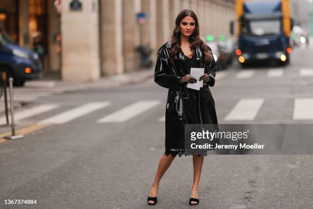 Gabrielle Caunesil wears white pearls in the hair, a black lace print pattern V-neck knees dress, a black shiny vinyl belted trench coat, black...