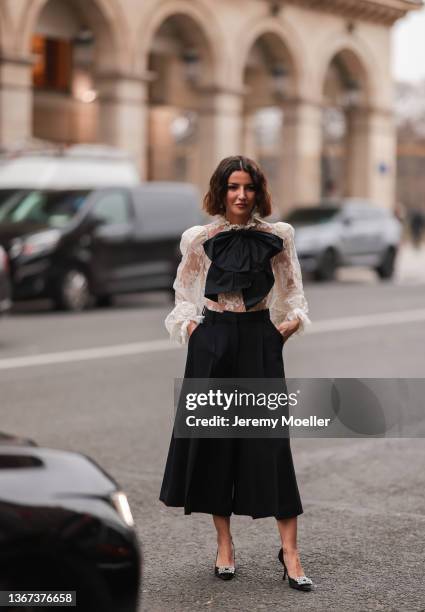 Alexandra Pereira wears large pearl pendant earrings, a white lace print pattern shoulder-pad shirt, a black oversized tie-bow, a black high waist...