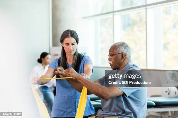mid adult physical therapist teaches senior man elastic band exercise - ergotherapie stockfoto's en -beelden