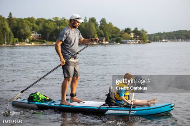 father and son stand-up paddleboarding on quiet lake during a warm sunny summer vacation day - sup stock pictures, royalty-free photos & images