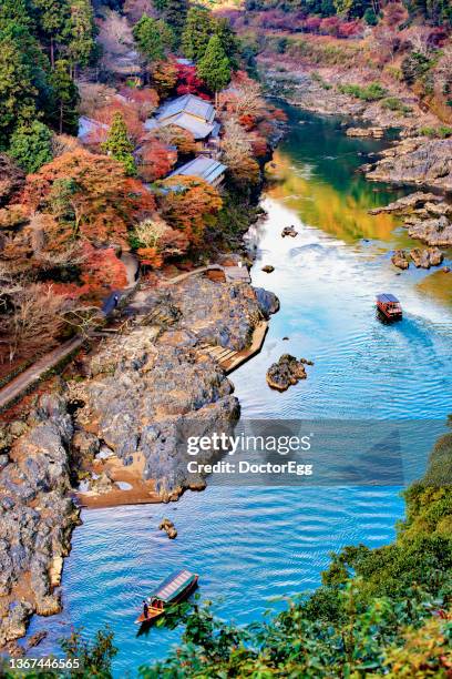 colourful maple trees on the hill with tourist boat in hozugawa river  from kameyama view point at arashiyama, kyoto, japn - autumnal forest trees japan stockfoto's en -beelden