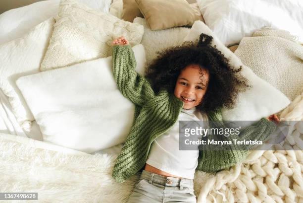 a little afro girl lies on a bed with many pillows. casual style. curly black hair. cute afro girl. - materasso foto e immagini stock