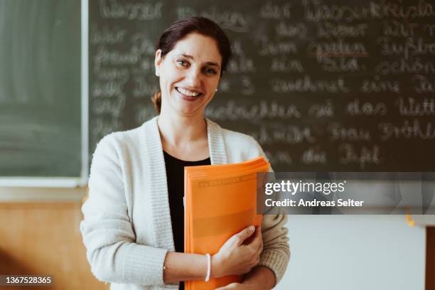 smiling female teacher in front of a chalkboard with exercise books in hands - teacher studio portrait stock pictures, royalty-free photos & images