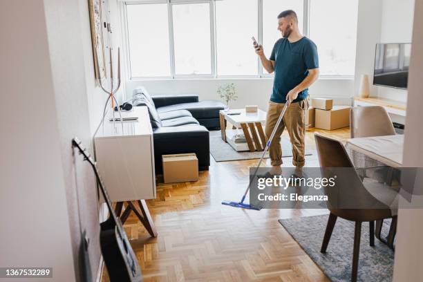 uomo che pulisce il suo appartamento e usa il suo smartphone, lava il pavimento del soggiorno, strumenti per la pulizia. straccio soffice in microfibra per il lavaggio. concetto di lavori domestici e pulizie di primavera - sweeping foto e immagini stock