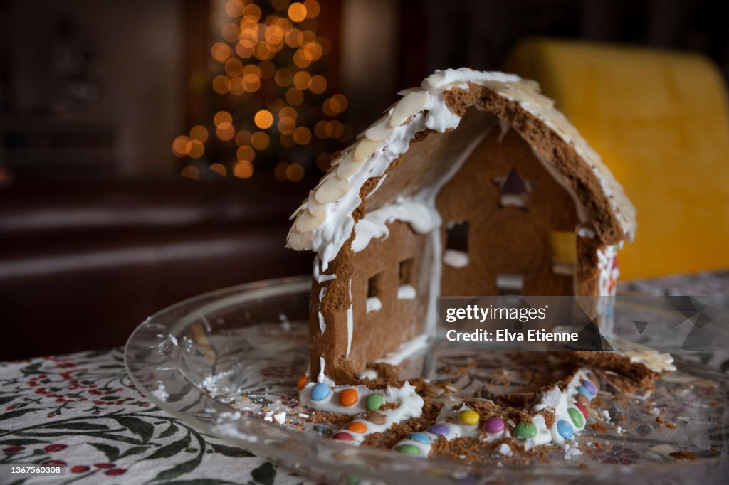 Half eaten Christmas gingerbread house decorated with icing and multi-coloured sweets on a glass plate on a dining table