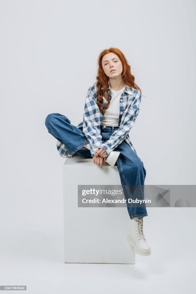 Beautiful young model posing sitting in studio on cube white background casual new clothes catalog. Fashion and trend concept