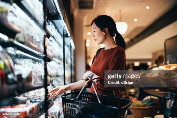 side profile of beautiful young asian woman carrying a shopping basket, grocery shopping for daily necessities in supermarket. healthy eating lifestyle. making healthier food choices - supermarket fruit stockfoto's en -beelden