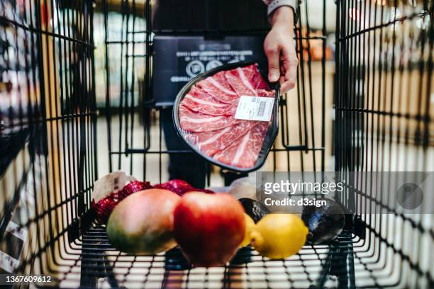 close up of woman pushing a shopping trolley, grocery shopping for fresh poultry in supermarket. she is putting a packet of steak into the shopping cart. routine grocery shopping. healthy eating lifestyle - cultivated meat stock pictures, royalty-free photos & images