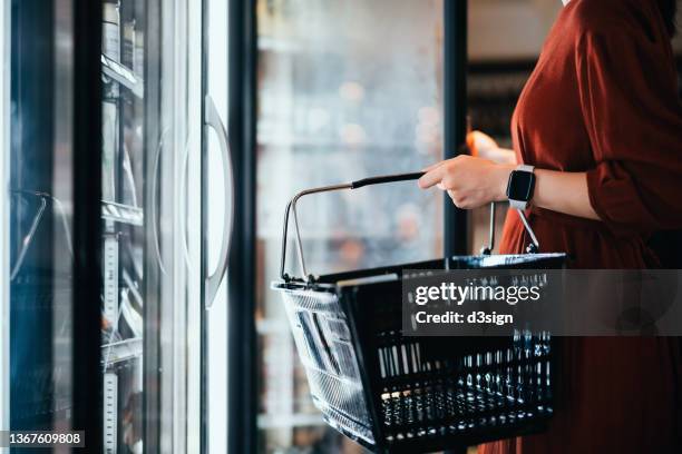 mid-section of young asian woman carrying a shopping basket, standing along the cold produce aisle, shopping for frozen food and daily necessities in supermarket. healthy eating lifestyle. making healthier food choices. home cooking lifestyle - freeze tag stock pictures, royalty-free photos & images