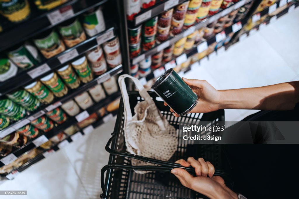 High angle view, close up hands of young Asian woman grocery shopping in supermarket. She is putting a tin can into a cotton mesh eco bag in a shopping basket. Environmentally friendly and zero waste concept