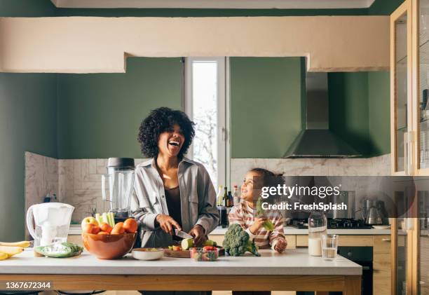 an african-american single mother preparing vegan lunch in the kitchen and smiling with her little daughter - baby eating food imagens e fotografias de stock