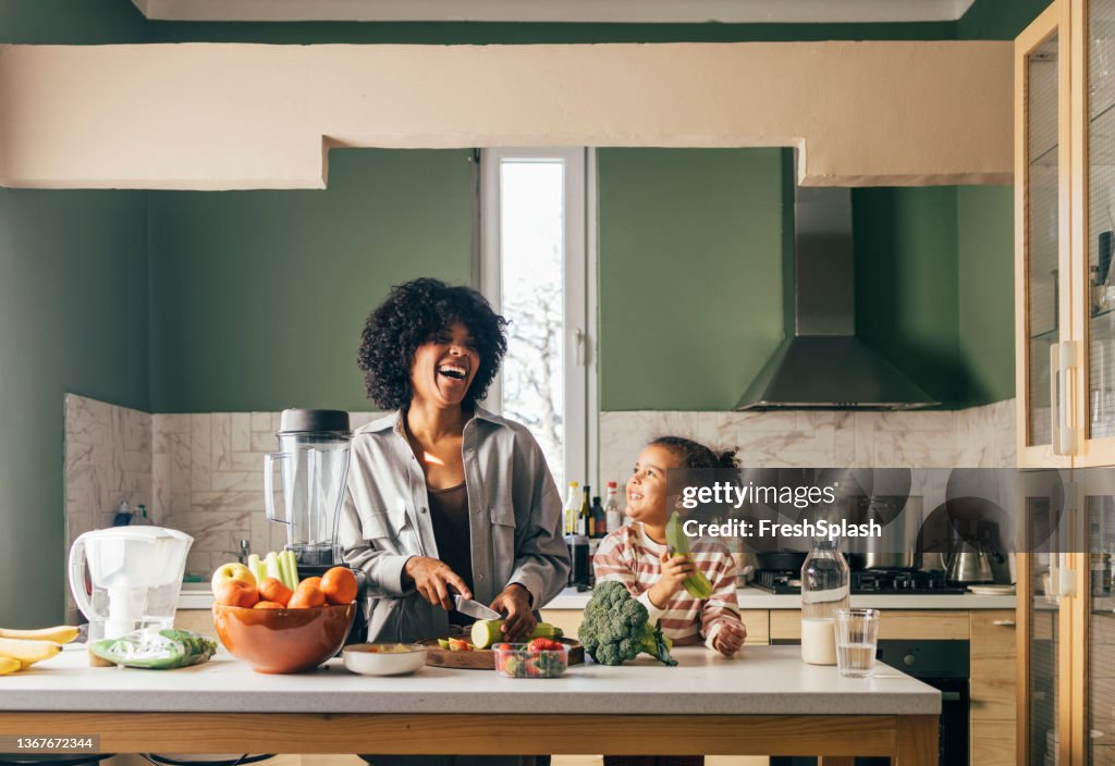 An African-American Single Mother Preparing Vegan Lunch In The Kitchen And Smiling With Her Little Daughter