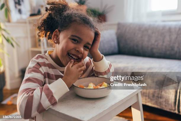 a portrait of a cute african-american girl sitting in her living room smiling and enjoying eating an orange - funny black girl stock pictures, royalty-free photos & images