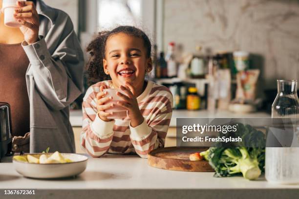 ein wunderschönes afroamerikanisches mädchen, das lächelt, während sie den smoothie trinkt, den ihre unkenntliche mutter für sie gemacht hat - yellow milk stock-fotos und bilder