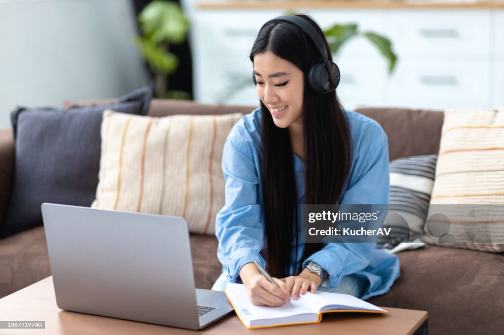 Smiling pretty Asian girl in headphones sitting at sofa, writes in a notebook and looking at laptop screen. Online work, education at home, e-learning
