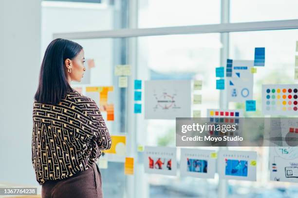 foto retrovisora de una joven diseñadora haciendo una lluvia de ideas con notas en una pared de vidrio en una oficina - publicidad fotografías e imágenes de stock