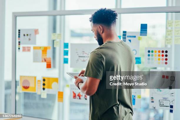 foto retrovisor de un joven diseñador usando una tableta digital mientras hace una lluvia de ideas con notas en una pared de vidrio en una oficina - publicidad fotografías e imágenes de stock