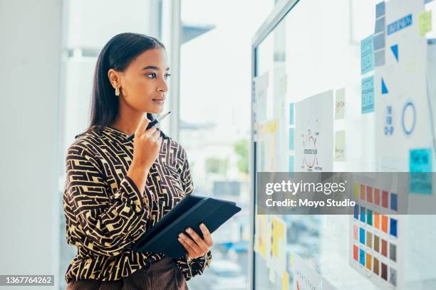 shot of a young designer using a digital tablet while brainstorming with notes on a glass wall in an office - advertentie stockfoto's en -beelden