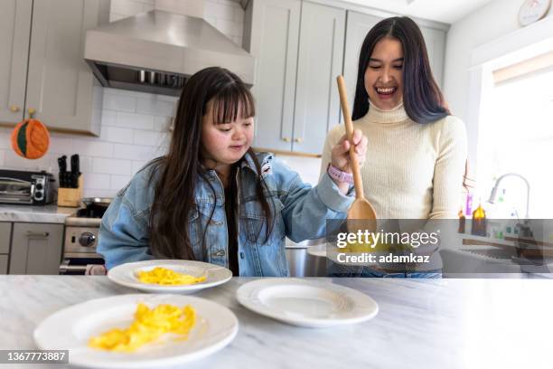 sisters bringing plates of eggs to the breakfast table. one sister has down's syndrome. - down syndrome cooking stock pictures, royalty-free photos & images