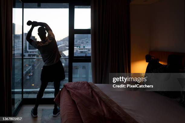 young man stretching by bedroom window looking at city view at sunrise - warm up exercise stock pictures, royalty-free photos & images