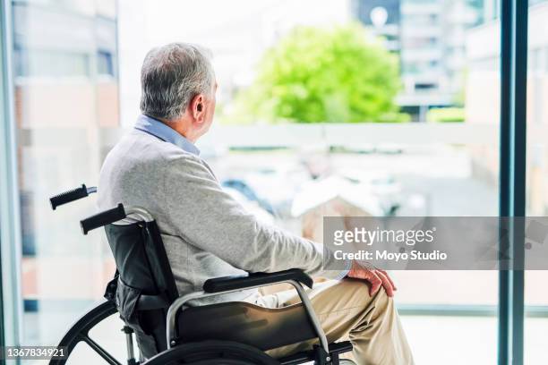 shot of a senior man looking out of a window while sitting in a wheelchair at a clinic - indecisive stockfoto's en -beelden