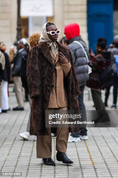 Guest wears gold rings in the hair, white sunglasses, a beige and white print pattern silk scarf, a beige turtleneck pullover, a brown oversized...