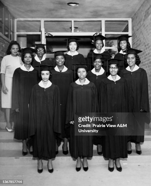 Eleven female students dressed in cap and gown for graduation and one female in a dress posing for the camera at Prairie View A&M University, 1960s