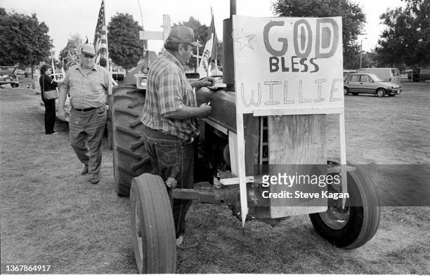 View of an unidentified man as he stands beside a tractor the features a handwritten sign at the debut Farm Aid benefit festival, Champaign,...