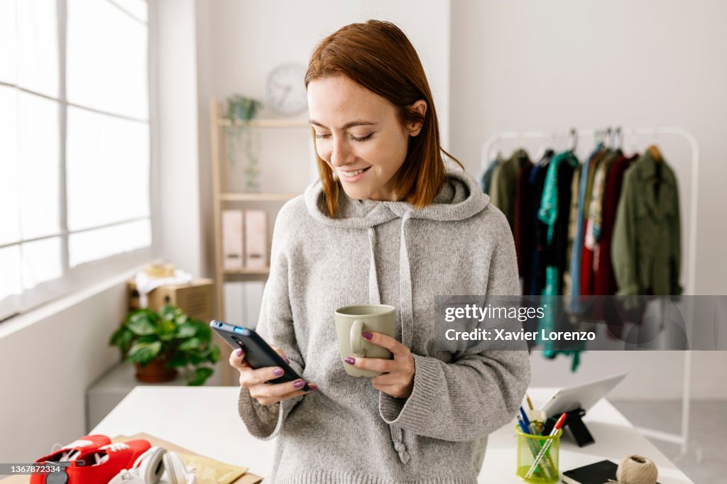 Female owner using cell phone and drinking coffee while standing in home office