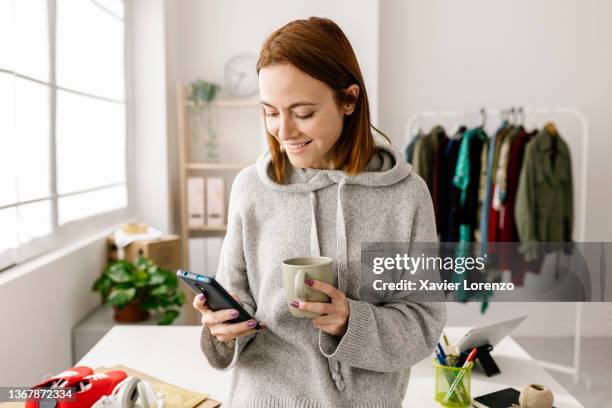 female owner using cell phone and drinking coffee while standing in home office - coffee shop stockfoto's en -beelden