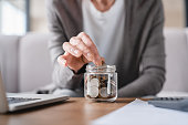 Cropped focused shot. Savings concept. Nest egg of old elderly senior woman grandmother saving money, economizing pension, mortgage loan at home using laptop and putting coin into moneybox