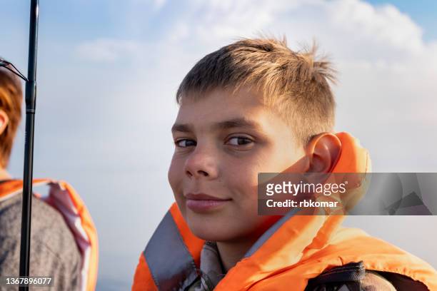 portrait of a happy tween boy in a bright orange life jacket looking at the camera during summer fishing on the lake. safety of children on the water, personal rescue equipment - deep sea fishing stock pictures, royalty-free photos & images