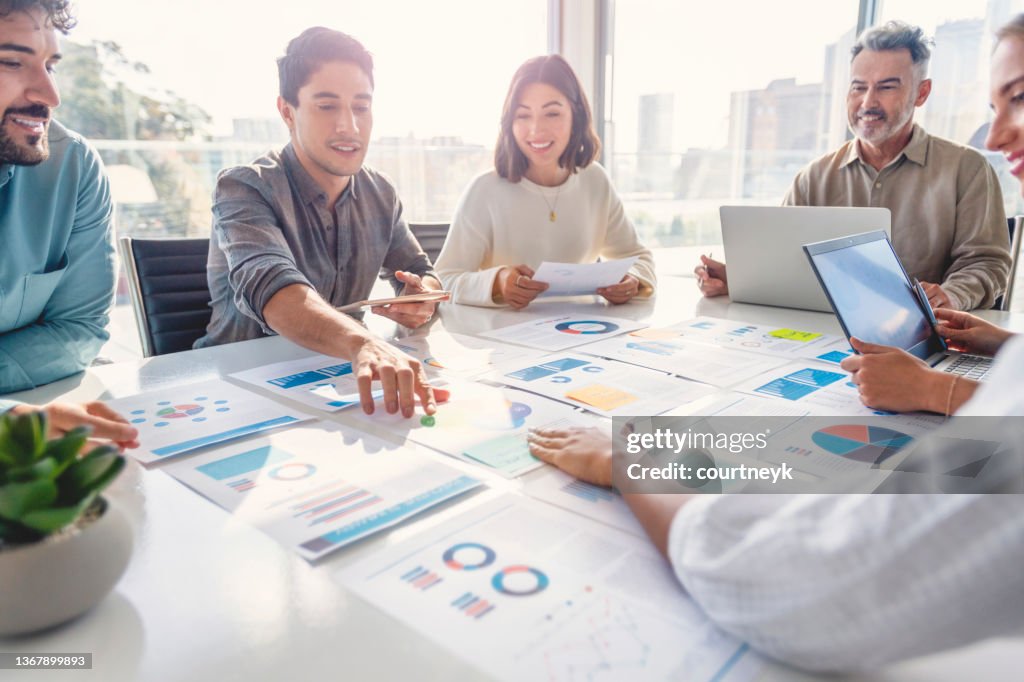 Multirassisch vielfältige Gruppe von Menschen, die mit Papierkram auf einem Sitzungstisch bei einer Geschäftspräsentation oder einem Seminar arbeiten. Die Dokumente enthalten Finanz- oder Marketingzahlen, Grafiken und Diagramme. Auf dem Tisch liegen Lap