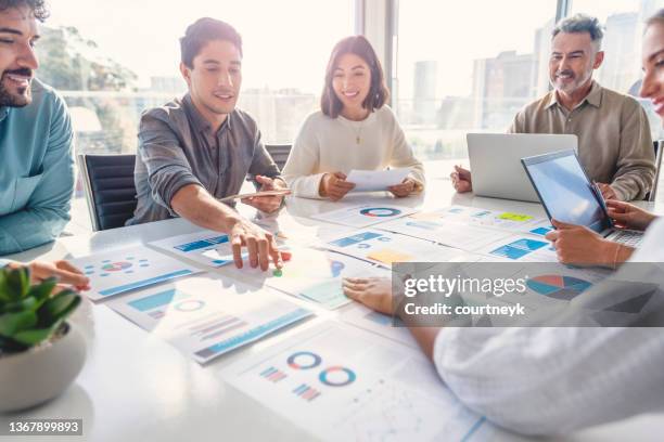 multi racial diverse group of people working with paperwork on a board room table at a business presentation or seminar. the documents have financial or marketing figures, graphs and charts on them. there are laptops and digital tablets on the table - communicate stockfoto's en -beelden