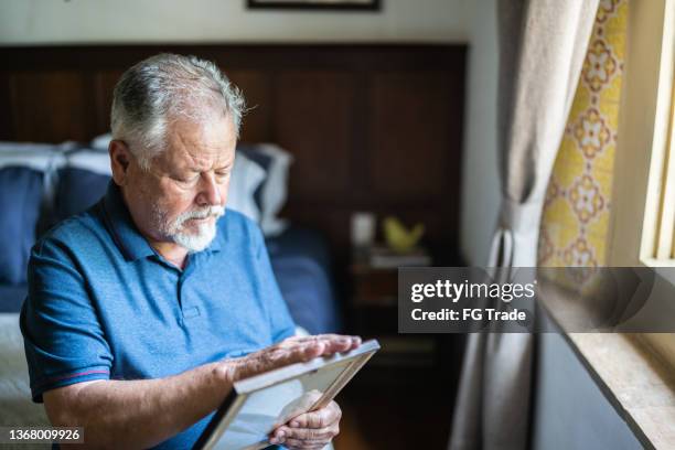 senior man holding a picture frame missing someone at home - mourner stockfoto's en -beelden