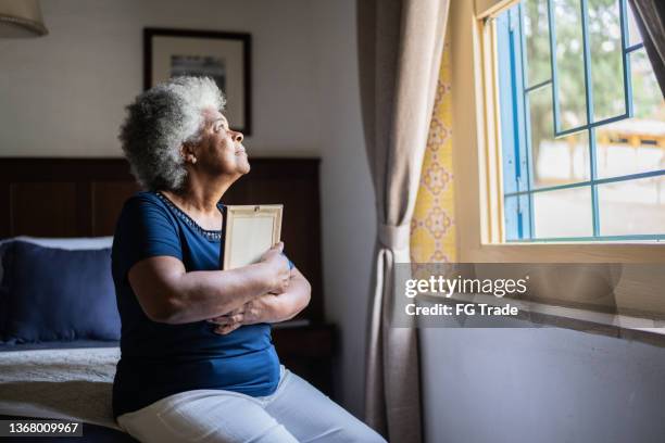 mujer mayor sosteniendo un marco de fotos que extraña a alguien en casa - viuda fotografías e imágenes de stock