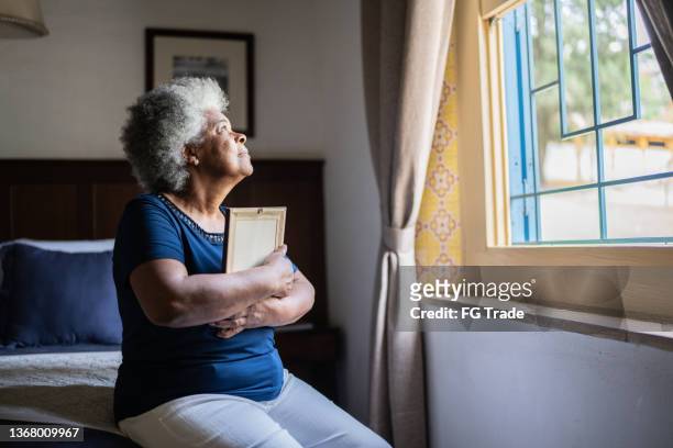 senior woman holding a picture frame missing someone at home - mourner stock pictures, royalty-free photos & images