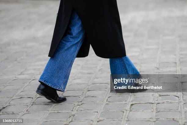 Guest wears a black long coat, blue denim flared pants, black shiny leather ankle shoes, outside Chanel, during Paris Fashion Week - Haute Couture...