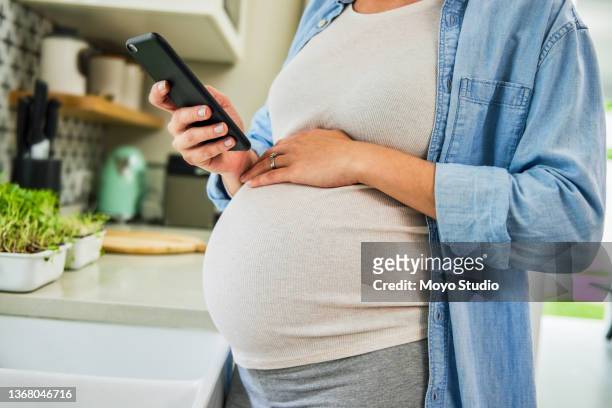 cropped shot of an unrecognisable pregnant woman standing alone in her kitchen and using her cellphone - pregnancy class stockfoto's en -beelden