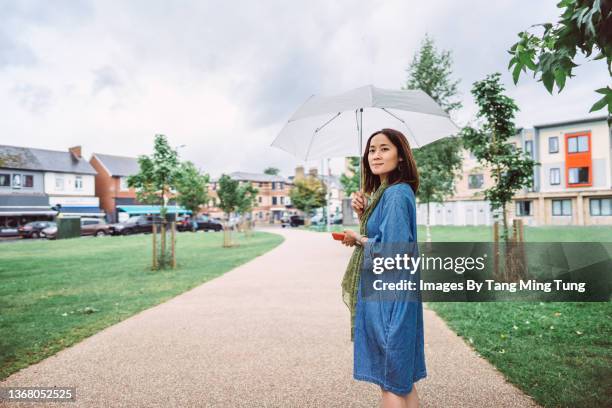 young pretty asian woman holding an umbrella and a smart phone while walking in the park during rain - female with umbrella stock-fotos und bilder