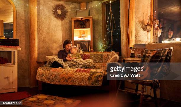 madre e hijo sentados en el sofá dentro de una cabaña ecológica por la noche - winter family home fotografías e imágenes de stock